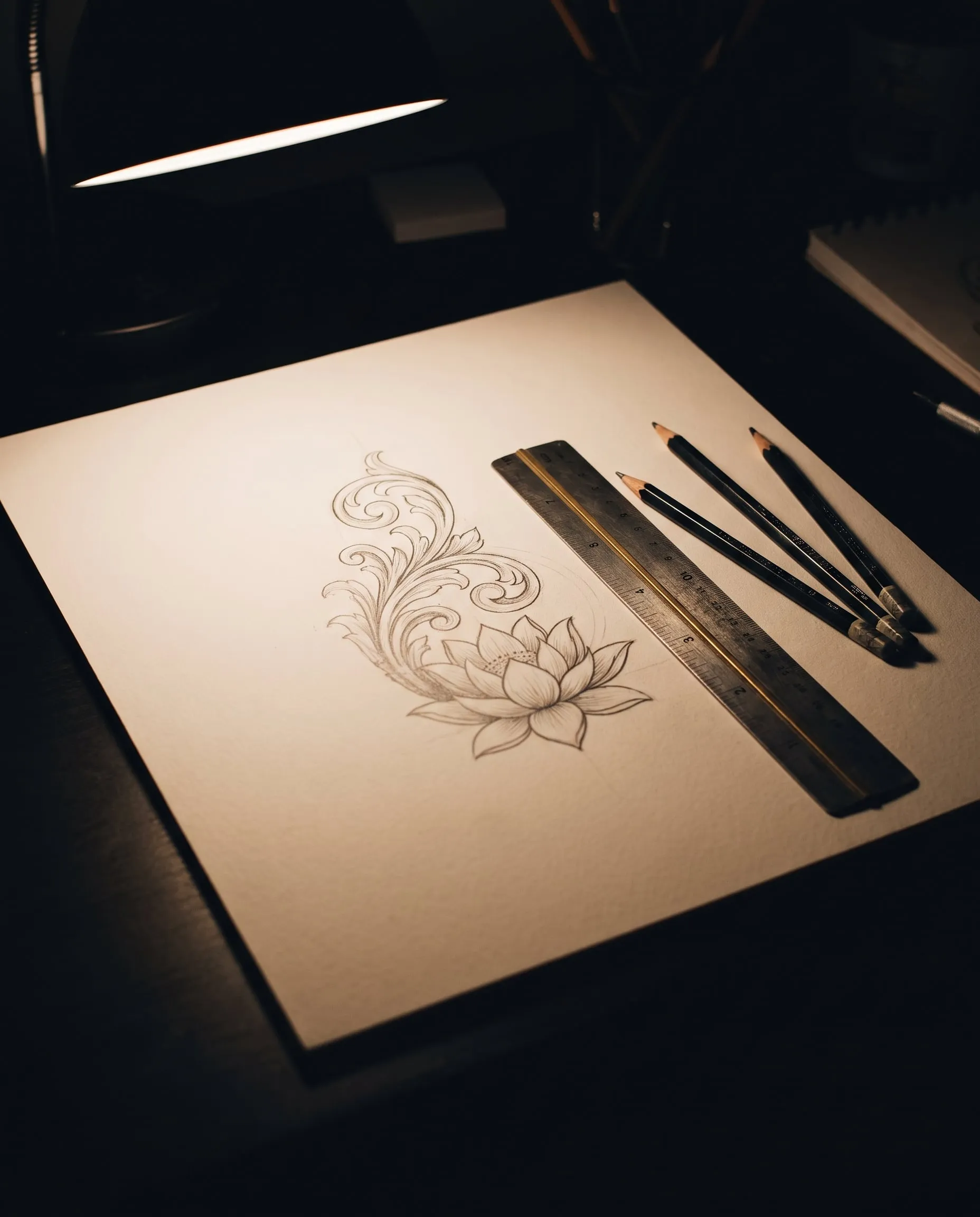 A graphite drawing of a lotus and scrollwork beside a steel ruler and pencils under a desk lamp