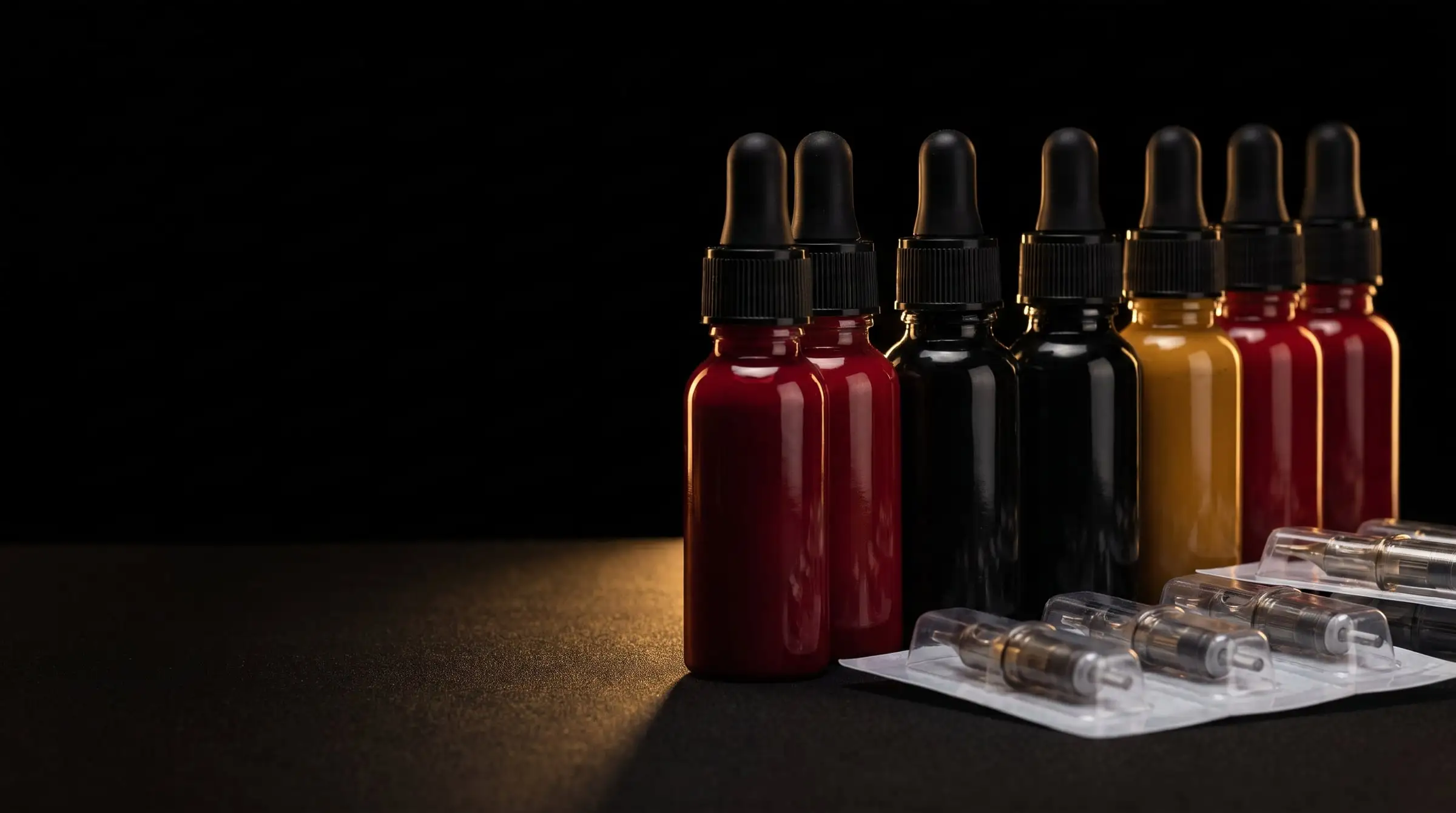 A row of red, black and gold tattoo ink bottles beside sealed needle cartridges