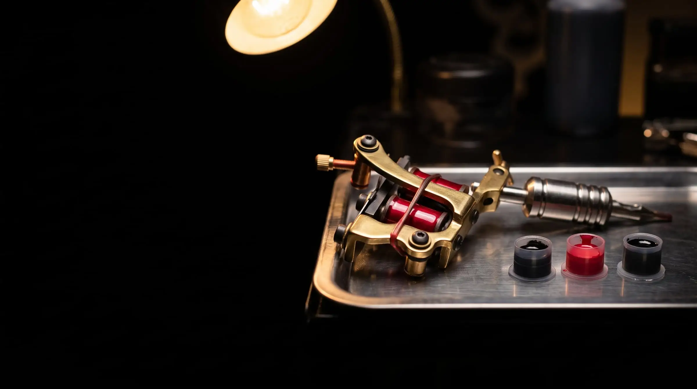 A brass coil tattoo machine with red coils on a steel tray beside caps of black and red ink