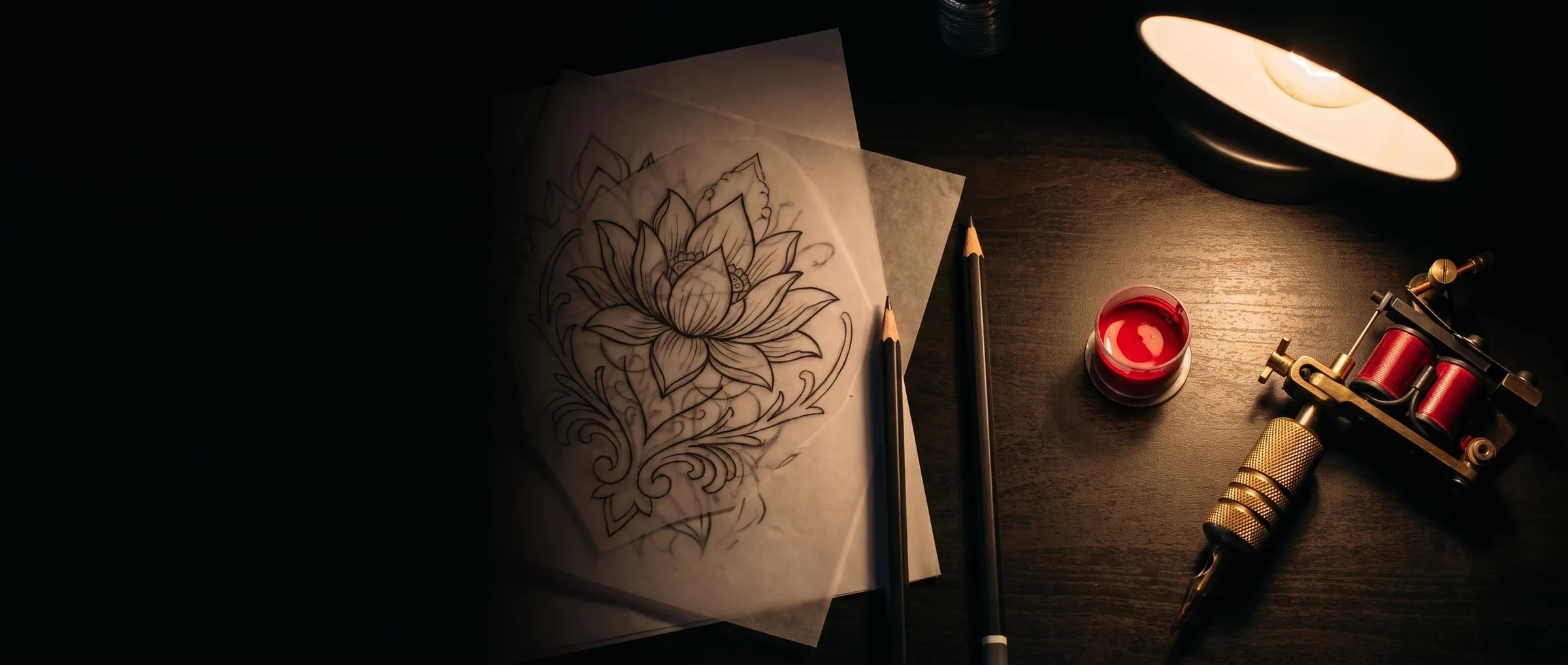 A dark desk with lotus linework on tracing paper, pencils, a cap of red ink and a coil tattoo machine