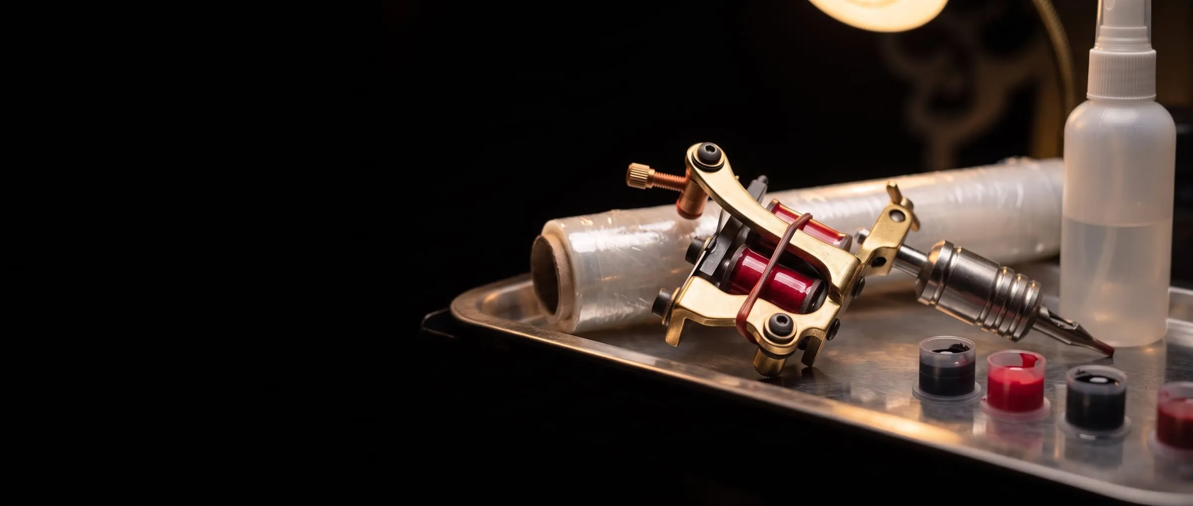 A coil tattoo machine, cling film, a spray bottle and ink caps on a steel tray in warm light