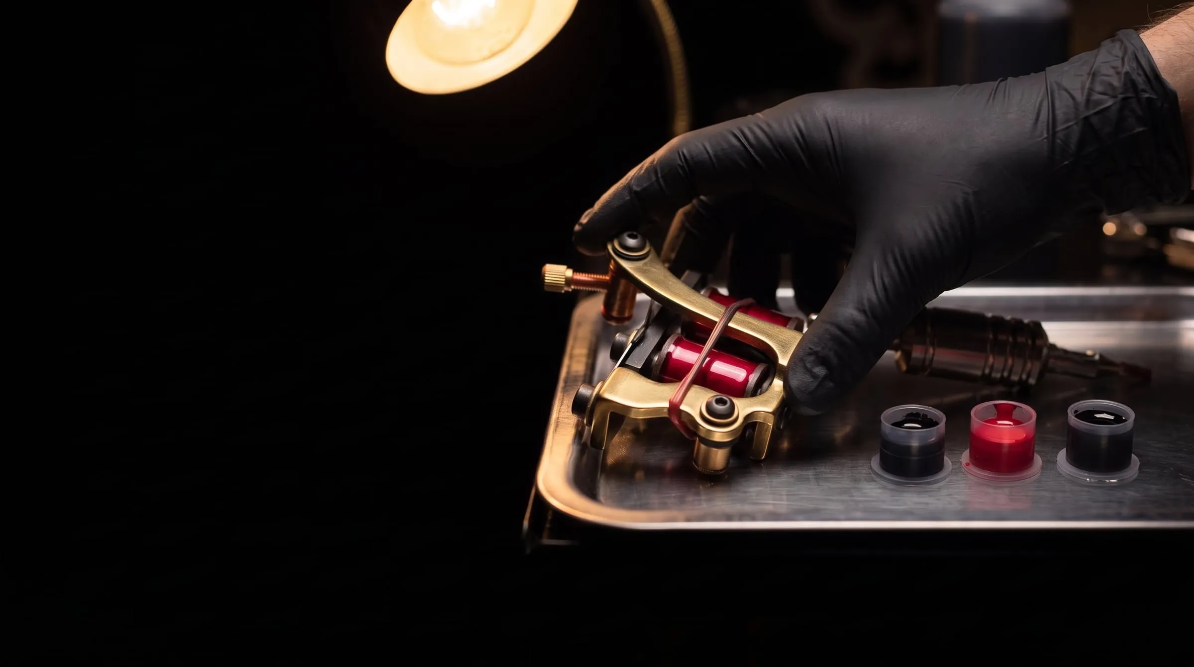 A hand in a black glove setting a coil tattoo machine down on a steel tray beside ink caps